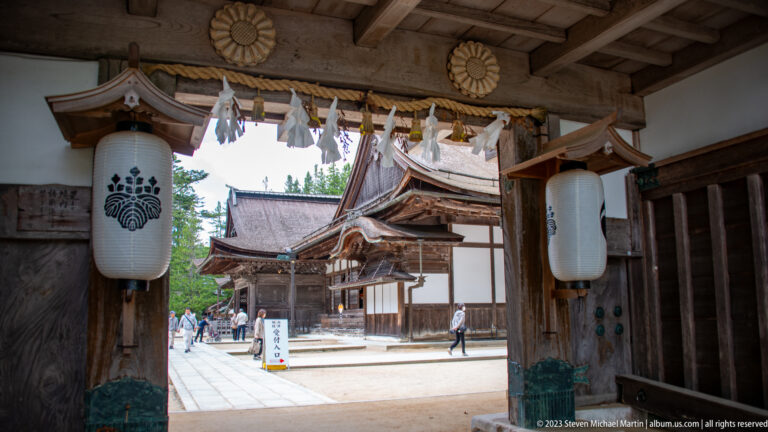 Timber temple gate with white crest lanterns and shimenawa framing traditional buildings inside the compound.
