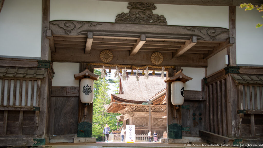 Frontal view through a timber gate with hanging lanterns, shimenawa, and a roofed temple hall beyond.