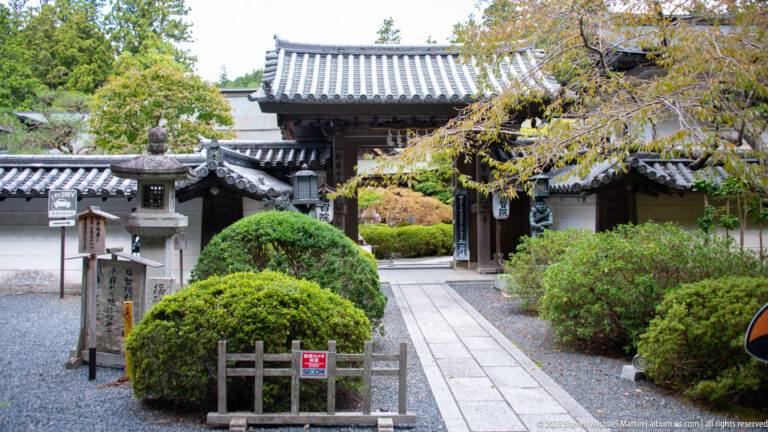 Stone path through a tiled gate bordered by rounded shrubs, gravel, and stone lanterns.