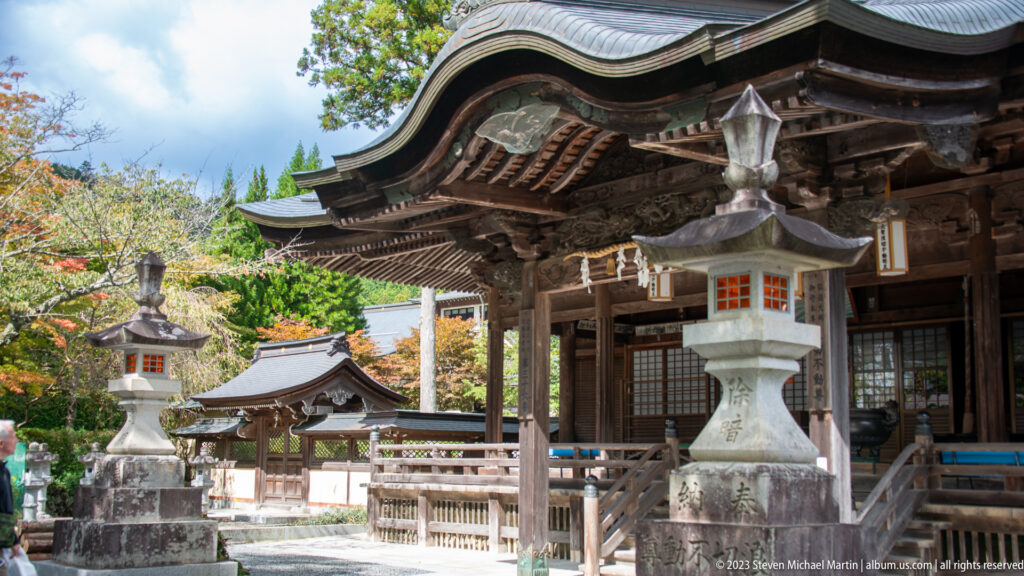Oblique view of a timber worship hall with curved roof, deep eaves, veranda, and stone lanterns.