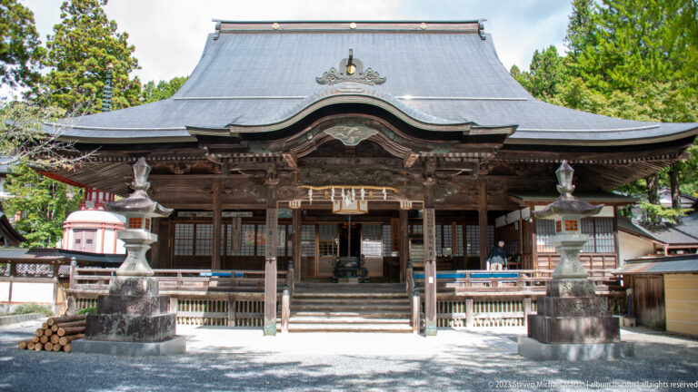 Frontal view of a Buddhist temple hall with curved roof, central steps, and paired stone lanterns.