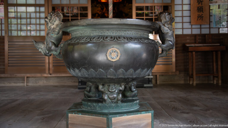 Large bronze incense burner with dragon handles, decorative bands, and a crest medallion before a temple hall.