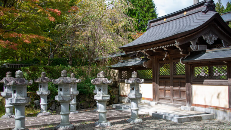 Row of stone lanterns beside a timber gate with curved tiled roof and white plaster walls.