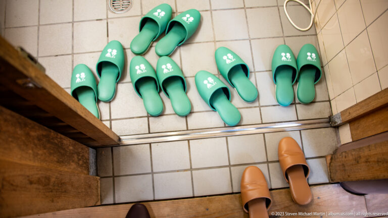 Green visitor slippers arranged on tiled flooring beside wooden steps and two brown indoor slippers.