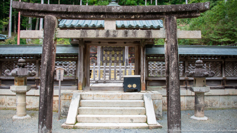 Stone torii before a small gated shrine enclosure with stone steps and flanking lanterns.
