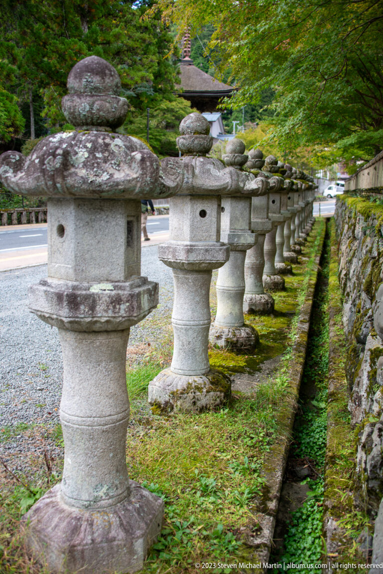 Long row of weathered stone lanterns receding beside a narrow drainage channel.