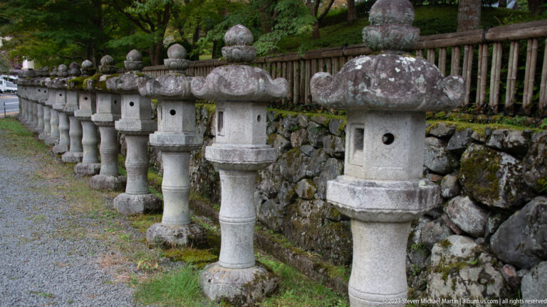 Close oblique view of weathered stone lanterns beside a low stone wall and wooden fence.