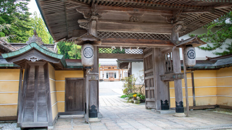 Interior view through a timber gate with crest lanterns, ocher walls, and stone paving beyond.
