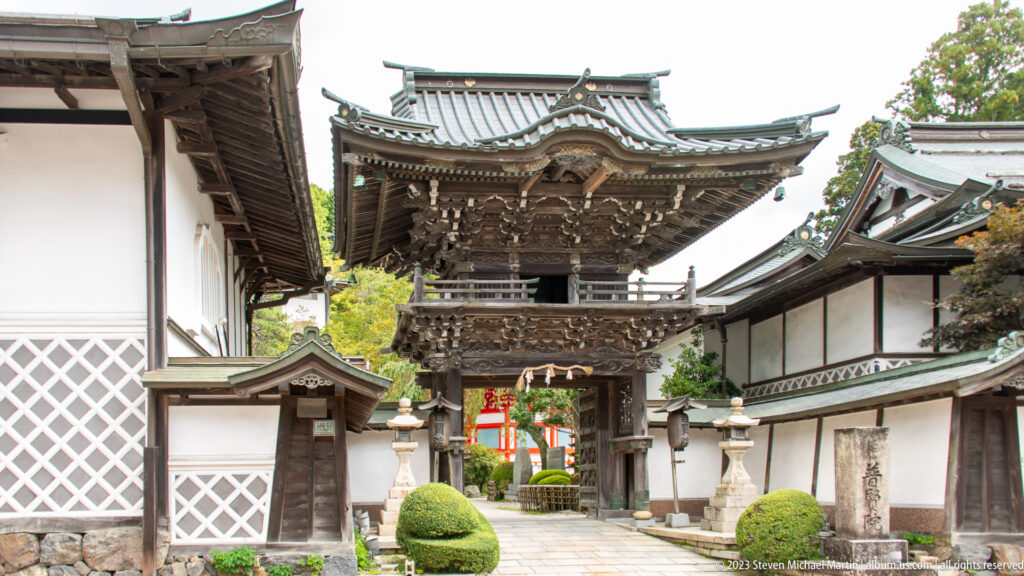 Two-story timber gate between white plaster walls with shrubs, lanterns, and vermilion architecture beyond.