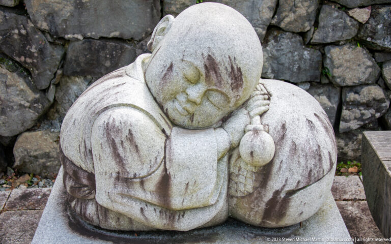 Small stone figure with closed eyes resting its head on folded hands beside a rough stone wall.