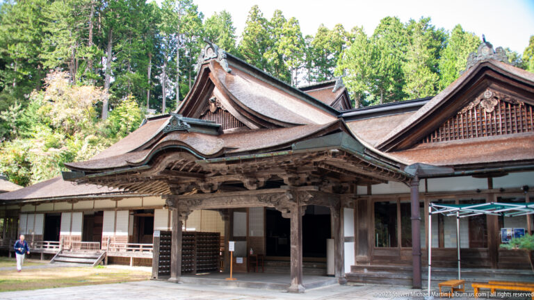 Traditional timber temple hall with curved karahafu entrance canopy, layered roofs, and white wall panels.