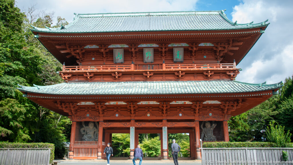 Vermilion two-story Daimon gate with tiled roofs and visitors walking through its central passage.
