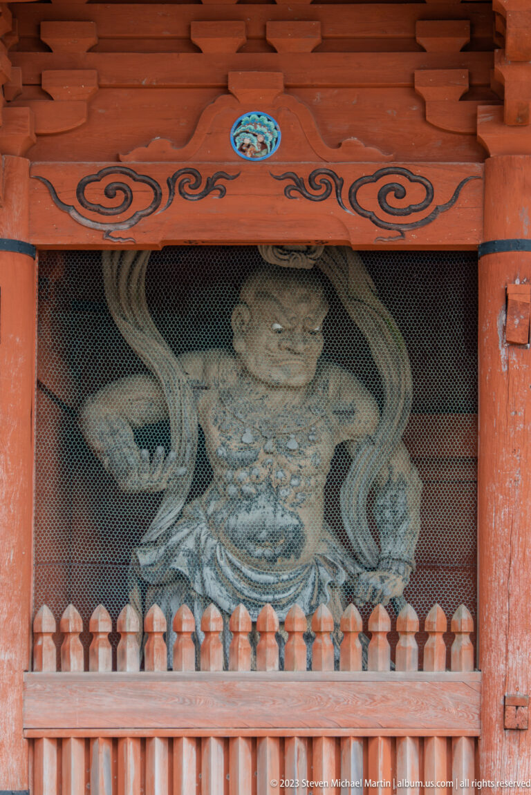 Large wooden Niō guardian behind mesh in a vermilion gate bay with painted cloud motifs.