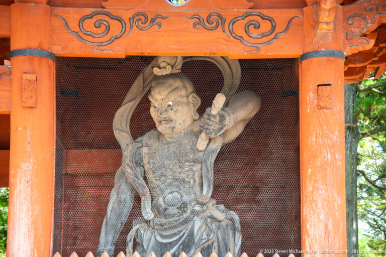 Wrathful wooden Niō guardian leaning forward behind mesh inside the Daimon gate.