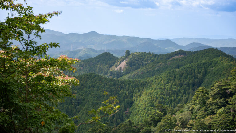 Layered forested mountain ridges with a pale cleared corridor crossing one slope.