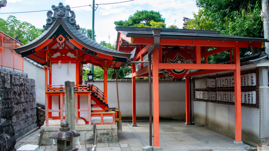 Small vermilion shrine and torii inside a narrow courtyard with inscribed offerings.