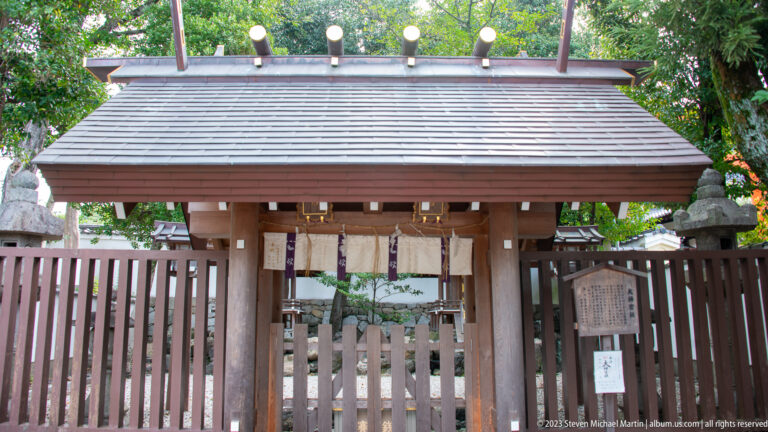 Wooden shrine gate with tiled roof leading into an enclosed sacred precinct.