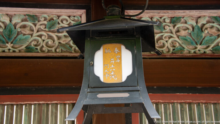 Metal shrine lantern with Japanese inscription beneath carved painted eaves.