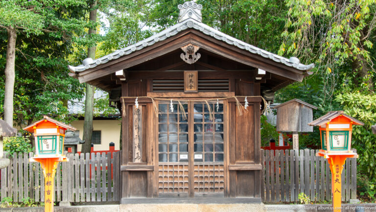 Small wooden shrine standing between two lanterns inside a fenced enclosure.