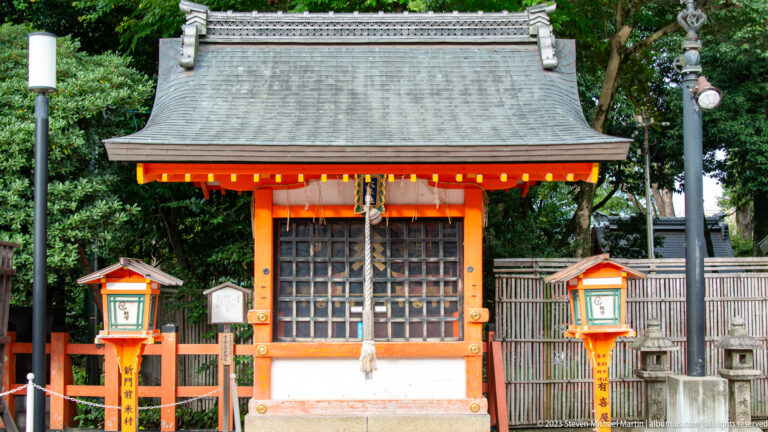 Small vermilion shrine with an offering area and paired lanterns.