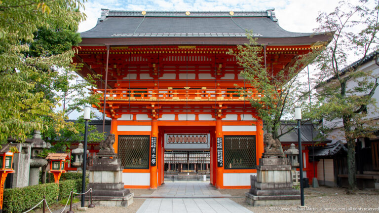 Two-story vermilion shrine gate with tiled roof and stone lanterns.