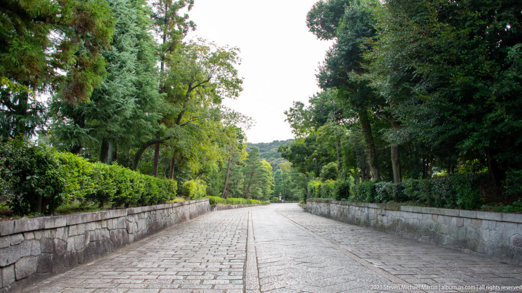 Long stone-paved path bordered by low walls, hedges, and mature trees.
