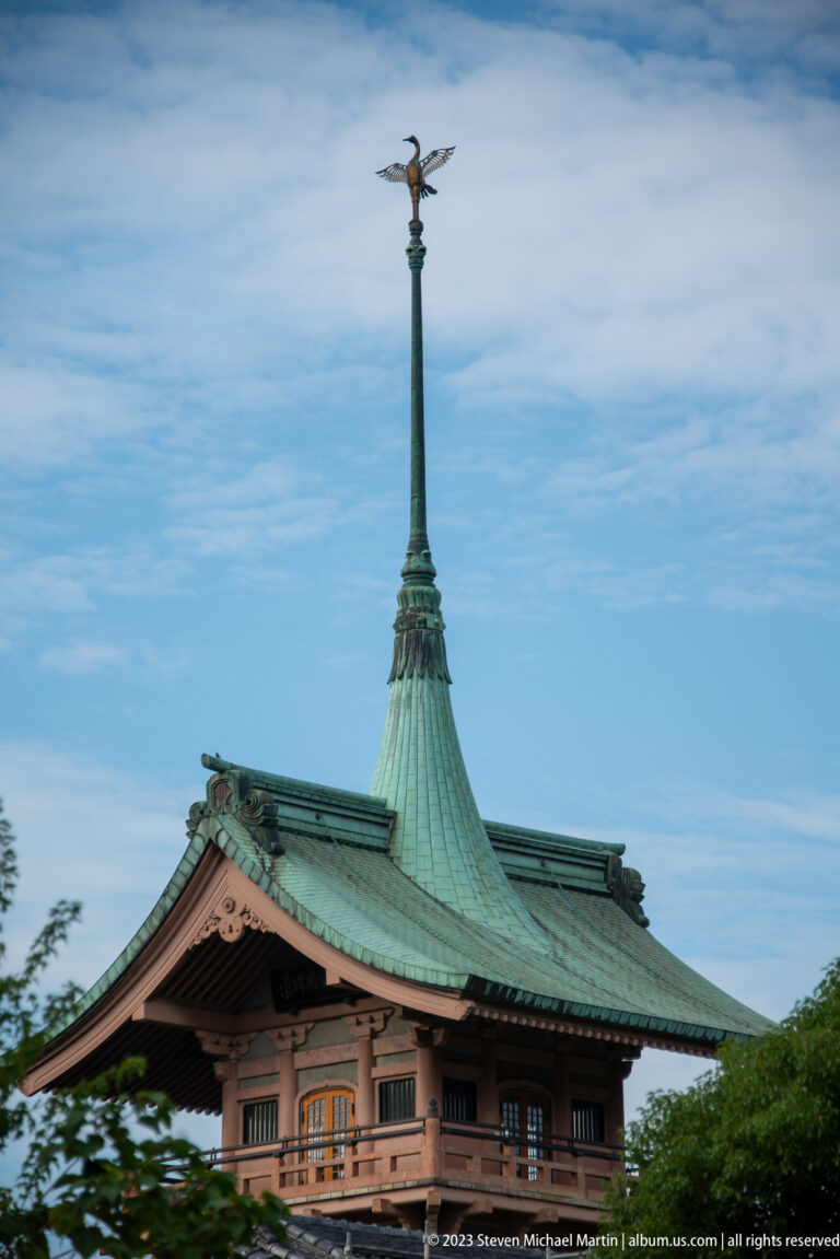 Green copper roof and tall spire of Gionkaku against blue sky.