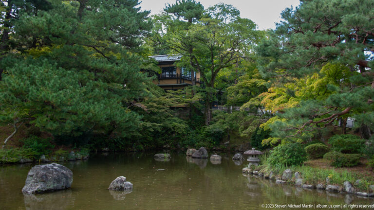 Landscaped pond with rocks, pine trees, stone lantern, and partly hidden pavilion.