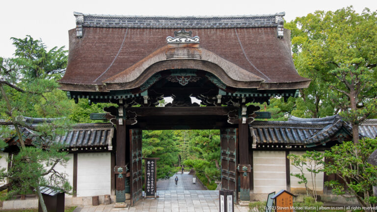 Main timber gate of Higashi Otani Sobyo opening onto a tree-lined path between white plaster walls.
