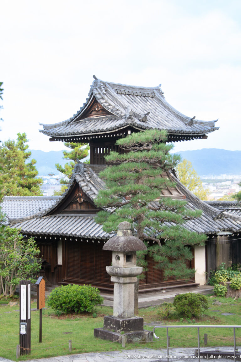 Open wooden pavilion with broad tiled roof, stone lantern, shrubs, and distant city view.