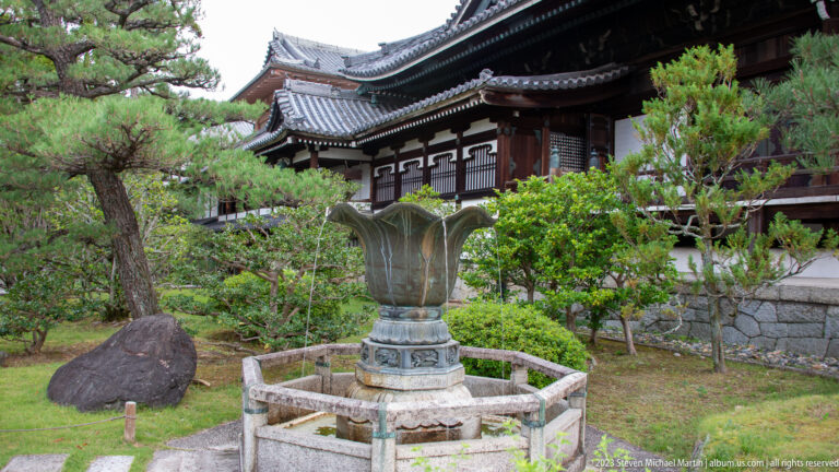 Large bronze lotus-shaped vessel on a stone base in a landscaped temple garden.