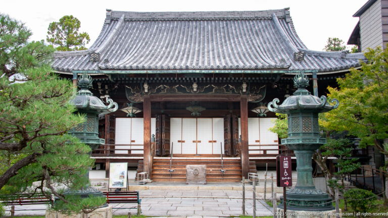 Temple hall with tiled roof, bronze lanterns, and stone forecourt in its surrounding Kyoto setting.