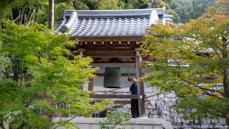 Bronze temple bell hanging beneath an open wooden bell tower in its surrounding Kyoto setting.