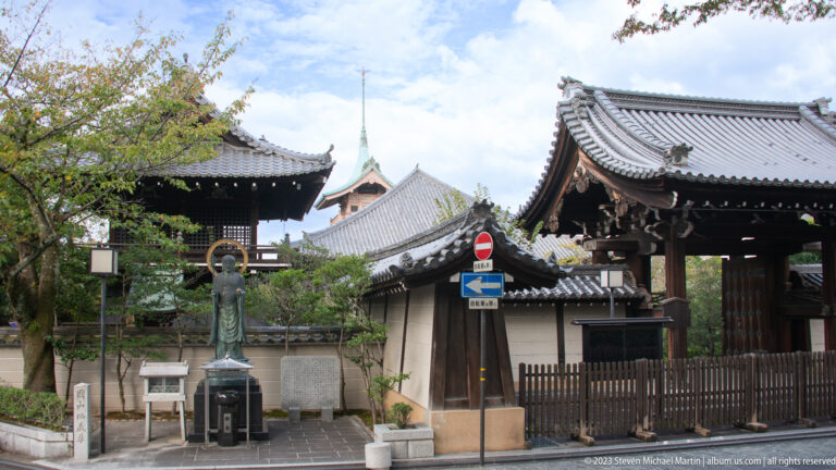 Standing Buddhist statue near a traditional temple gate and street in its surrounding Kyoto setting.