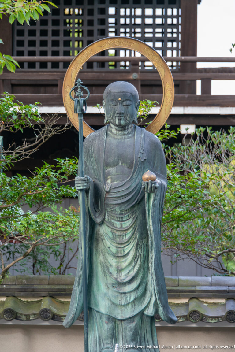 Bronze standing Buddhist statue with halo and staff in its surrounding Kyoto setting.
