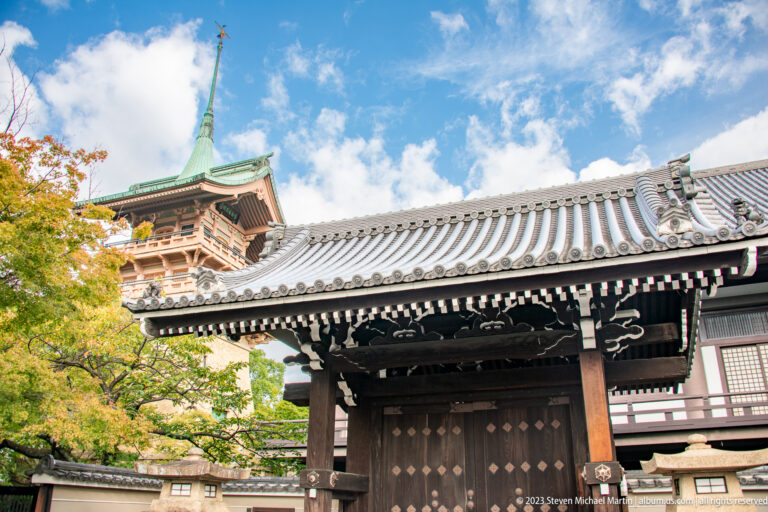 Timber temple gate with tiled roof and larger temple building behind in its surrounding Kyoto setting.