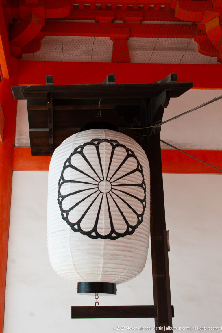 White paper lantern with black chrysanthemum crest hanging beneath vermilion beams at Heian Jingu.