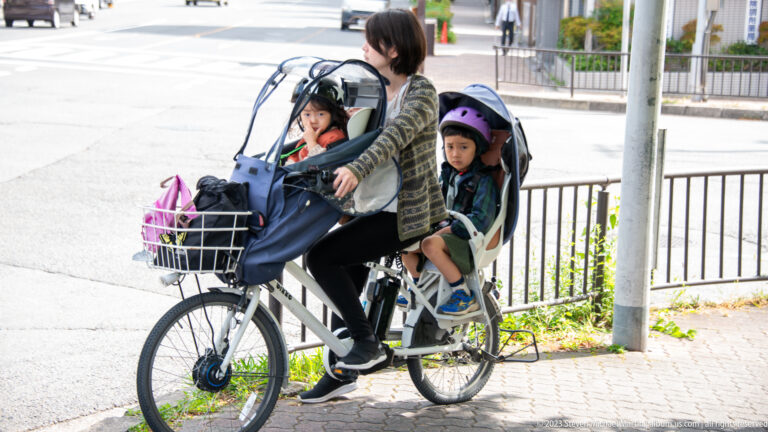 Adult riding bicycle with two children in child seats in its surrounding Kyoto setting.