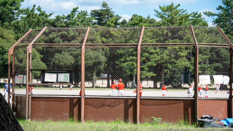Baseball players practicing behind tall fencing in its surrounding Kyoto setting.
