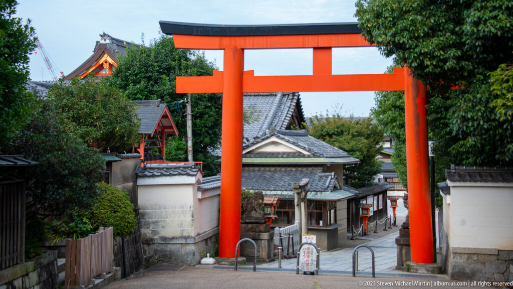 Large vermilion torii spanning a sloping lane lined with tiled roofs and shrine buildings.