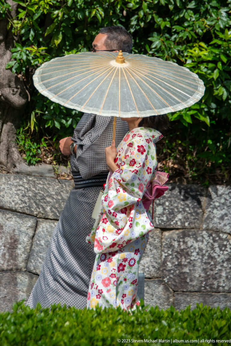 Adult and child in kimono under a paper umbrella in its surrounding Kyoto setting.