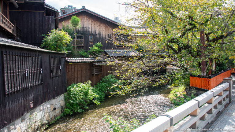 Shirakawa canal in Gion beside traditional wooden buildings, trees, and a low bridge.
