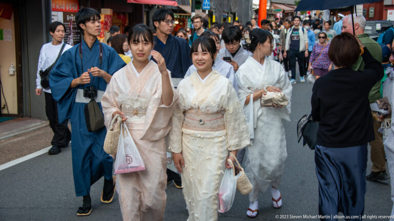 Crowd of visitors in kimono on a busy street in its surrounding Kyoto setting.