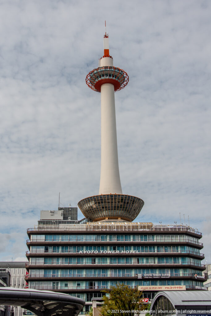 Kyoto Tower rising above the station district beneath a cloudy sky in its surrounding Kyoto setting.