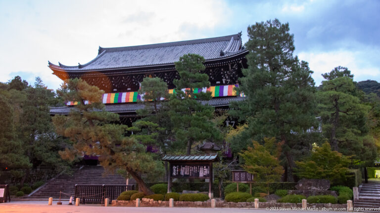 Large dark timber temple building behind trees, with a tiled roof and five-colored Buddhist cloths across the front.