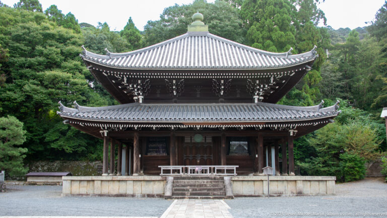 Symmetrical two-story dark wooden temple hall with tiled roofs, deep eaves, and a white-railed platform.