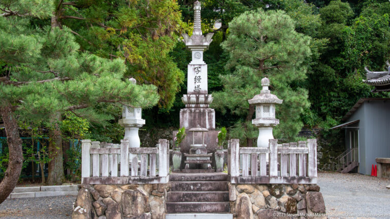 Tall inscribed stone memorial on steps, flanked by two stone lanterns and a low stone railing.