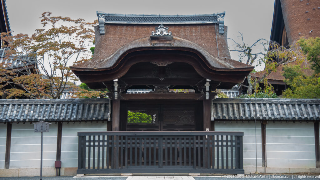 Ornate wooden gate with a curved karahafu gable between white plaster walls and gray tiled roofs.