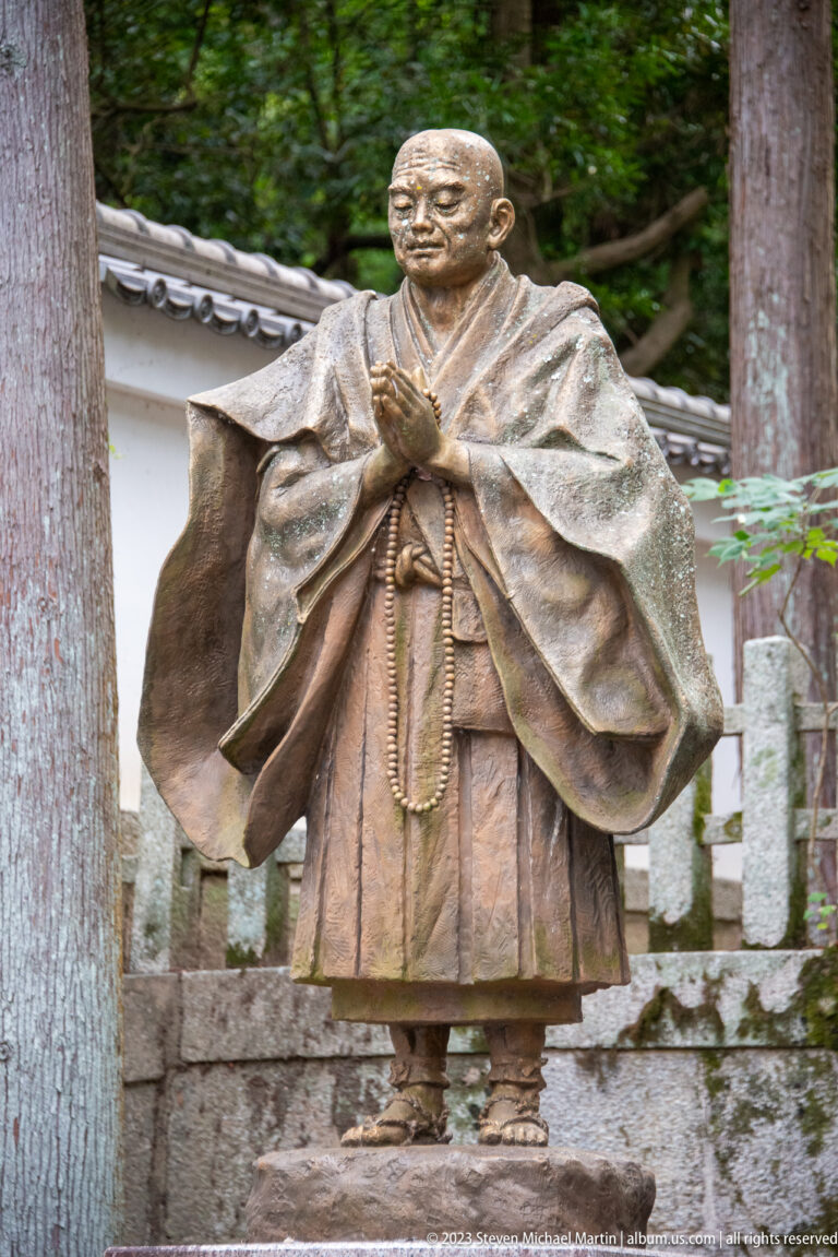 Close bronze statue of a standing Buddhist monk in layered robes holding long prayer beads.