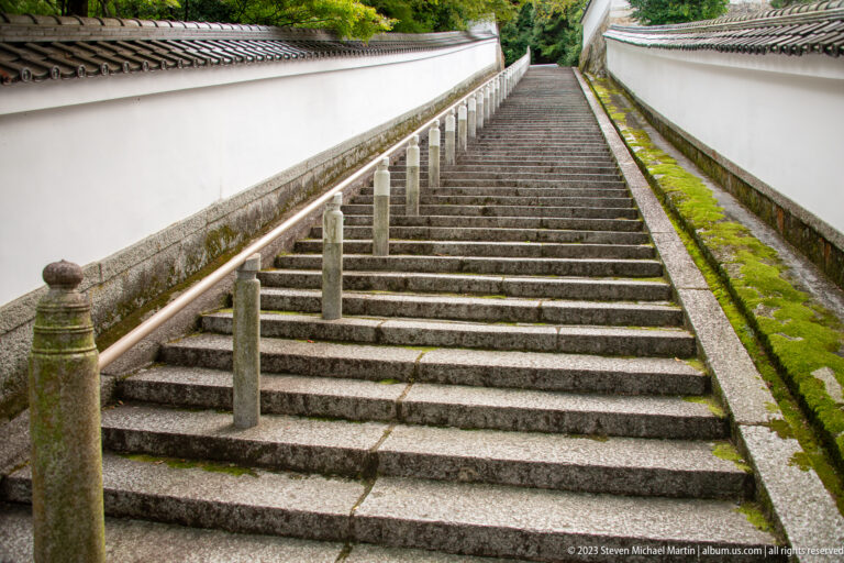 Long steep stone staircase rising between white plaster walls with low stone posts along the path.
